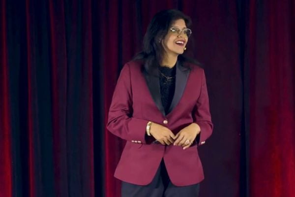 A portrait of Dr. Annie Kathuria, standing on stage with a red velvet curtain background, wearing a burgundy blazer with black lapels and glasses, smiling while looking to the right.
